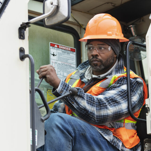A mature African-American man working at a construction site as a crane operator, sitting in the crane cab. He is wearing protective workwear - a hardhat, safety goggles, and reflective vest.