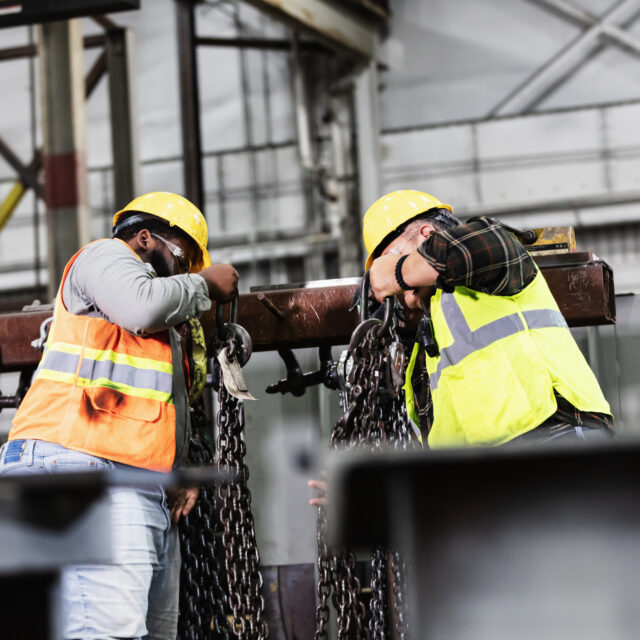 wo multiracial men working in a warehouse taking chain slings off a rack, to use with an overhead crane for lifting large, heavy objects. Their faces are mostly obscured by their hands holding the chains. The African-American man on the left is in his 20s. His Hispanic coworker is in his 50s.